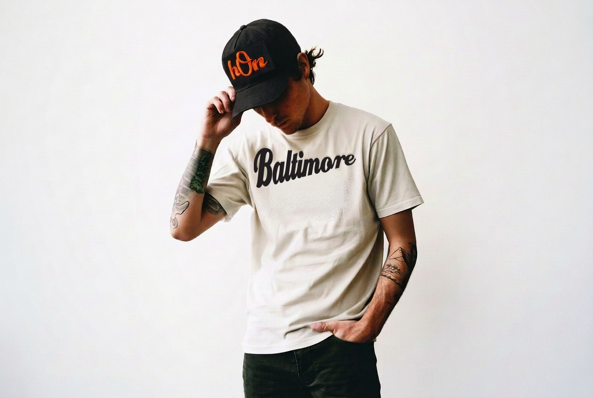 A man wearing a black baseball cap with orange embroidery and a light beige t-shirt with a subtle gray 'Allure' Baltimore logo, posing against a plain white background.